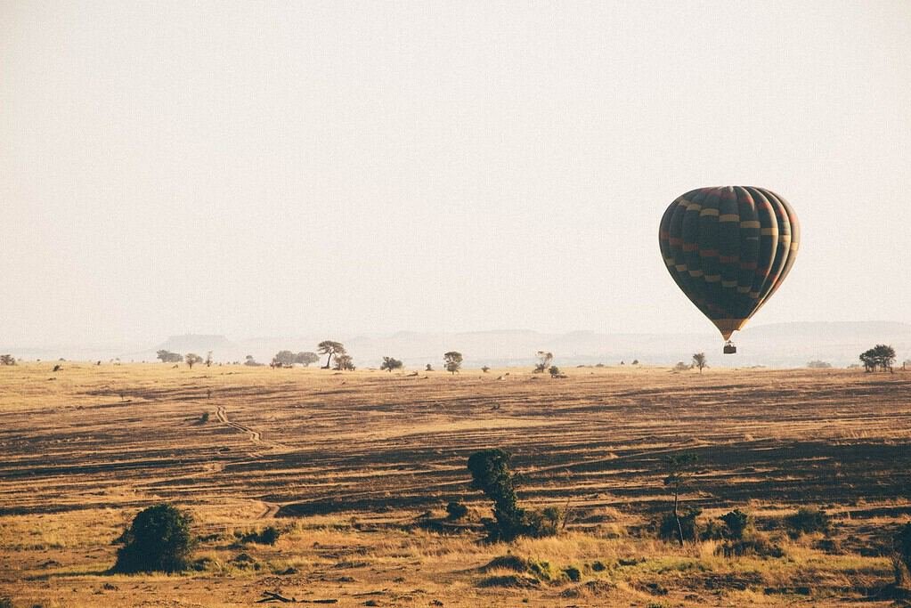 A hot-air balloon drifting over dry golden plains with scattered trees in hazy morning light