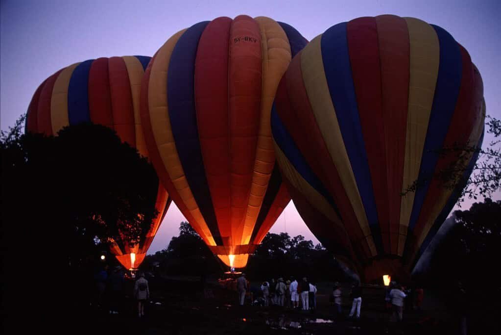About MaasaiMara.ke: plan and book your Maasai Mara safari 3 Three striped hot air balloons glowing as they inflate at dawn in Kenya, with safari guests waiting beside them