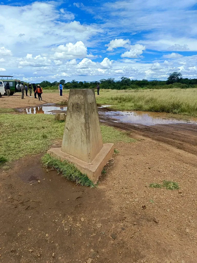 Maasai Mara entry fees: park fees, the 12-hour rule and how to pay 3 Concrete Kenya and Tanzania border pillar on a muddy track, with visitors and a safari vehicle behind