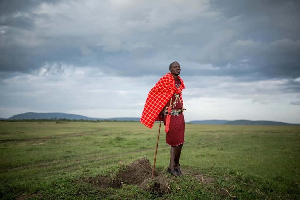 About Maasai Mara National Reserve 4 A Maasai guide in a red checked shuka leaning on his stick on open green plains under a cloudy sky