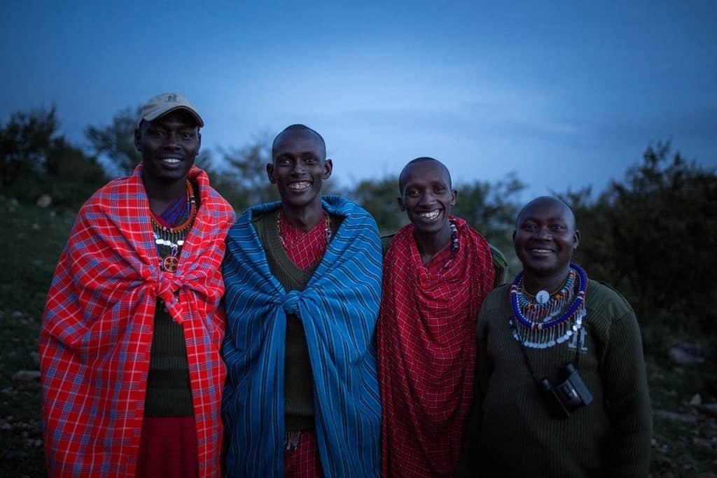 Four smiling Maasai safari guides and trackers in red and blue shukas, standing together at dusk