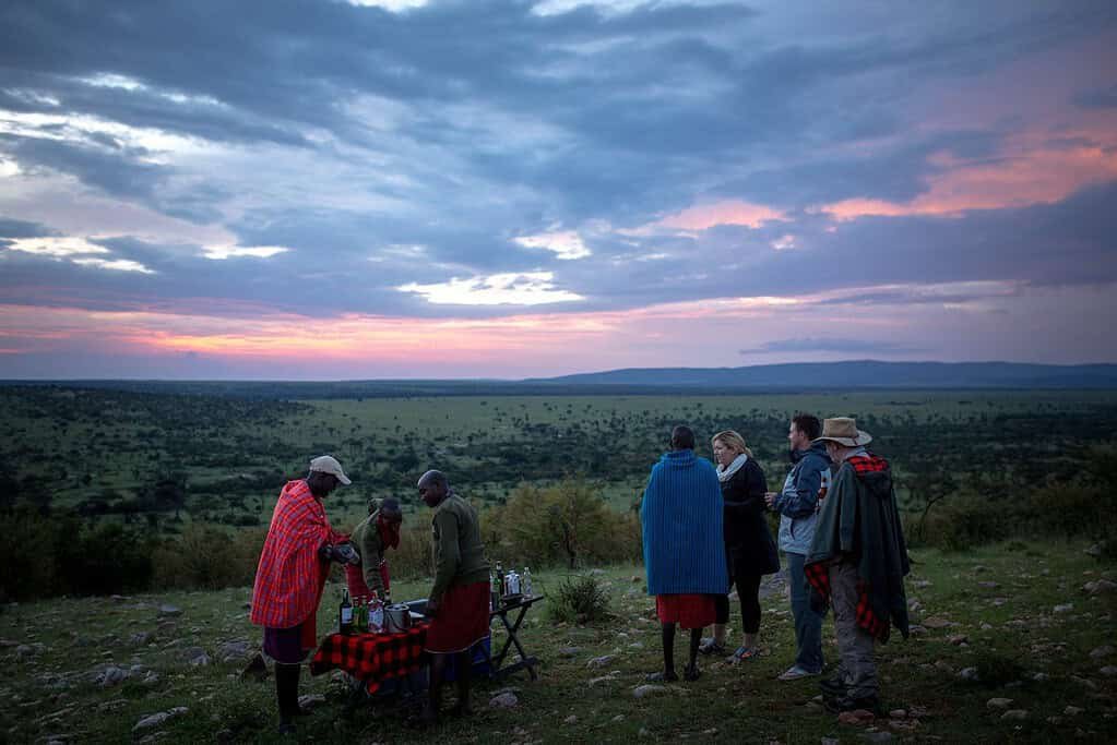 Maasai guides in red and blue shukas serving sundowner drinks to safari guests on a hilltop above the plains at dusk